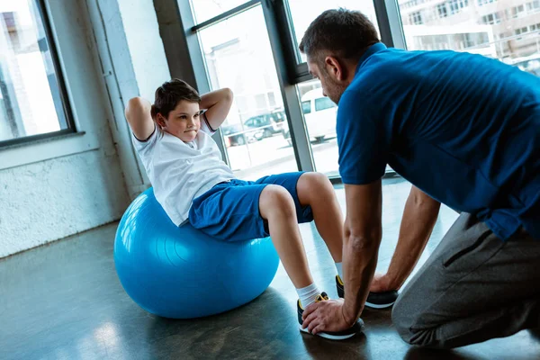 Father helping son sitting on fitness ball and doing sit up exercise at gym — Stock Photo