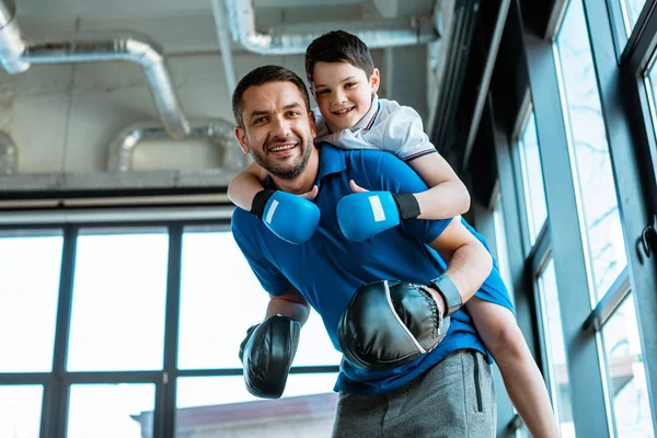 Happy father giving piggyback ride to happy son in boxing gloves at gym — Stock Photo