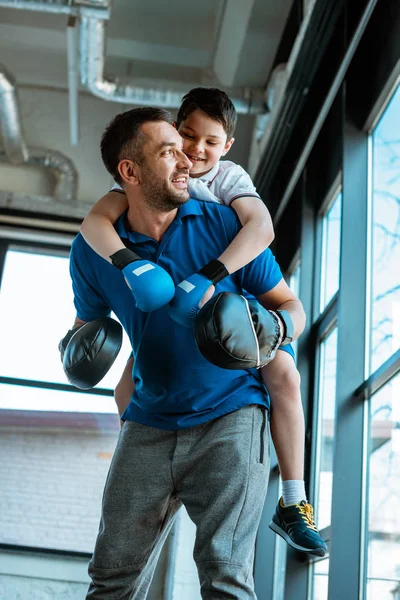 Happy father giving piggyback ride to smiling son in boxing gloves at gym — Stock Photo