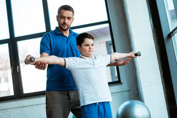 Pai ajudando filho a se exercitar com halteres no ginásio — Fotografia de Stock