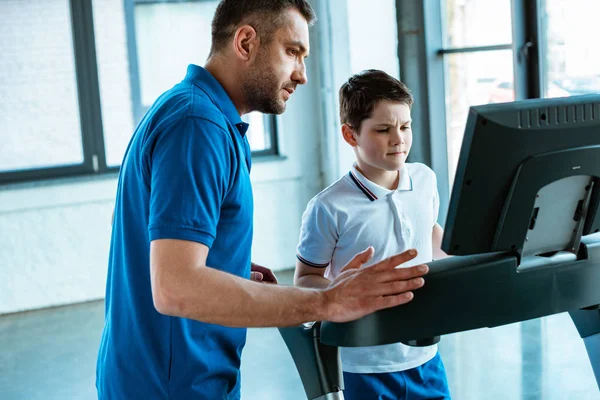 Vater schaut auf Bildschirm auf Laufband, während Sohn im Sportzentrum läuft — Stockfoto