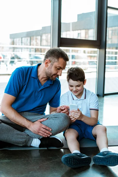 Father and son sitting on fitness mat and using smartphone at sports center — Stock Photo