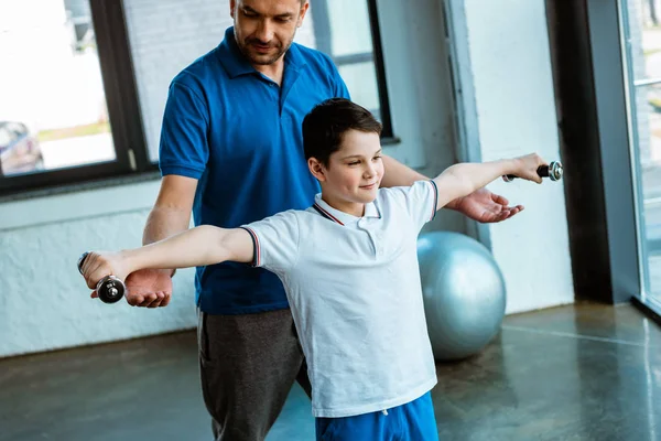 Padre ayudando a su hijo a hacer ejercicio con pesas en el centro deportivo - foto de stock
