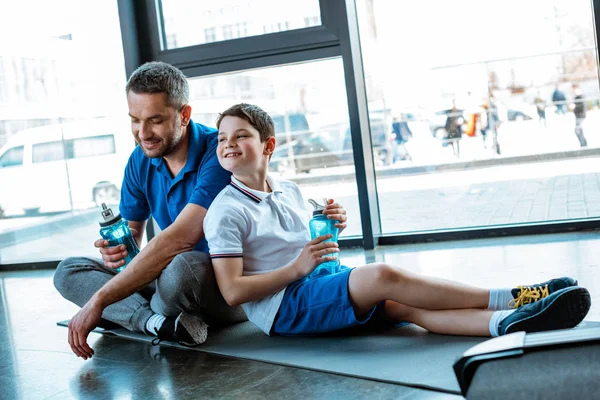 Sorrindo pai e filho sentado no tapete de fitness com garrafas de desporto no ginásio — Fotografia de Stock