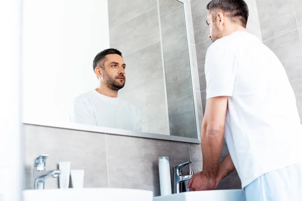 Красивый мужчина смотрит в зеркало и моет руки в ванной во время утренней рутины — стоковое фото