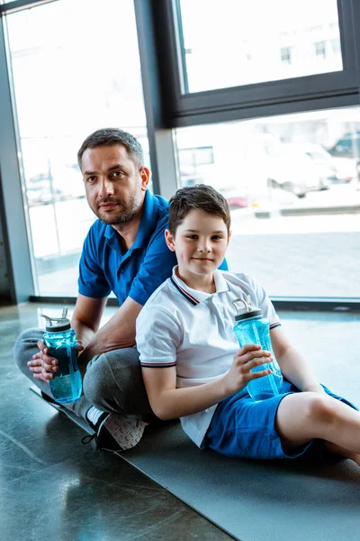 Padre e hijo sentados en la alfombra de fitness con botellas deportivas y mirando a la cámara en el gimnasio - foto de stock