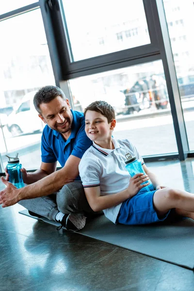 Feliz padre e hijo sentado en la alfombra de fitness con botellas de deporte en el gimnasio - foto de stock