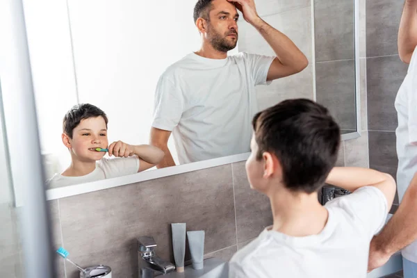 Син чистить зуби під час налаштування зачіски у ванній — стокове фото