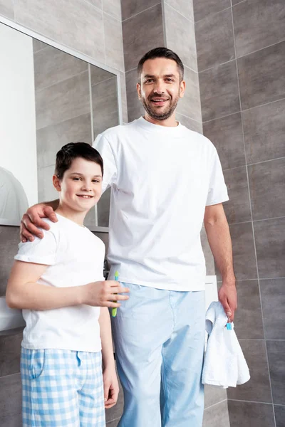 Счастливые отец и сын с зубными щетками смотреть в камеру в ванной комнате — стоковое фото