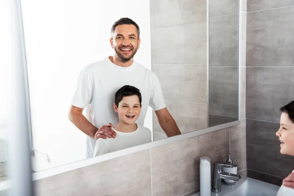 Vater blickt in die Kamera und umarmt lächelnden Sohn im Badezimmer während der Morgenroutine — Stockfoto