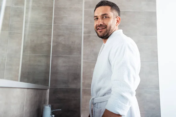 Красивый мужчина в белом халате смотрит на камеру в ванной комнате с копировальным пространством — стоковое фото