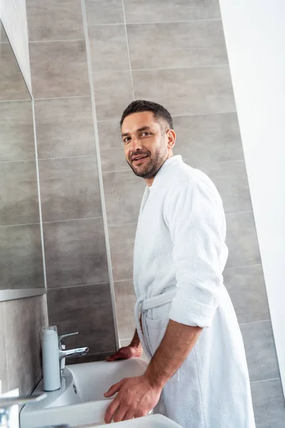 Hombre sonriente guapo en albornoz blanco mirando a la cámara en el baño durante la rutina de la mañana - foto de stock