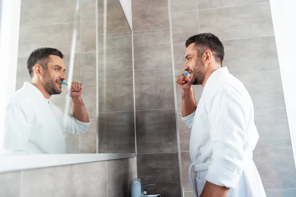 Hombre guapo de albornoz blanco cepillarse los dientes en el baño durante la rutina de la mañana - foto de stock