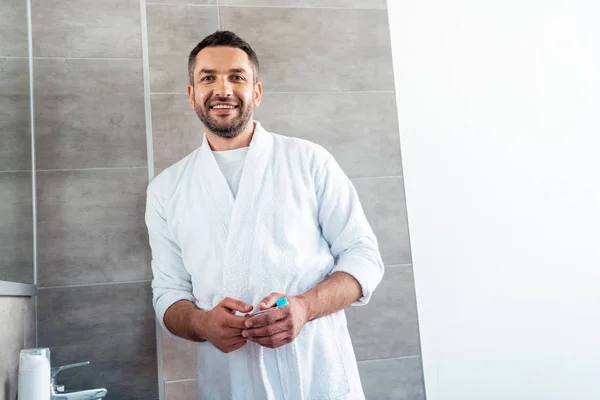 Красивий усміхнений чоловік у білому халаті з зубною щіткою дивиться на камеру у ванній — стокове фото