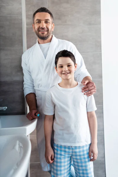 Усміхнений батько і син в піжамі дивиться на камеру у ванній — стокове фото