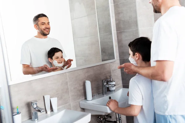 Padre aplicando crema de afeitar en la cara del hijo en el baño - foto de stock