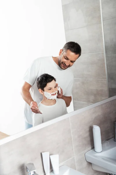 Sorrindo pai aplicando creme de barbear no rosto do filho no banheiro — Fotografia de Stock