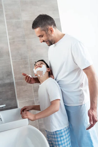 Pai aplicando creme de barbear no rosto do filho no banheiro durante a rotina da manhã — Fotografia de Stock