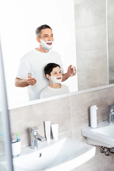 Padre feliz aplicación de crema de afeitar en la cara del hijo en el baño por la mañana - foto de stock