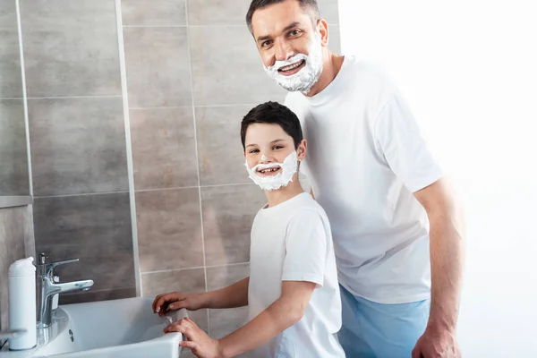 Sonriente hijo y padre con crema de afeitar en las caras mirando a la cámara en el baño - foto de stock