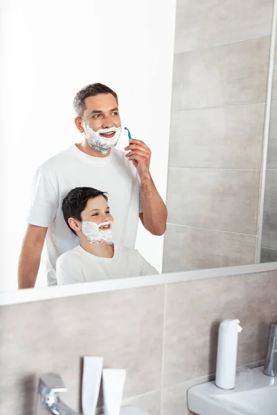 Son with shaving cream on face near father shaving with razor in bathroom — Stock Photo