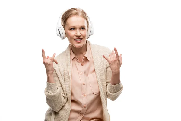 Mulher de meia idade em fones de ouvido ouvindo música e mostrando sinais de rock isolado em branco — Fotografia de Stock