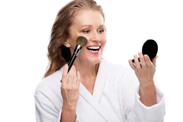 Femme mature en peignoir en utilisant miroir et pinceau cosmétique tout en appliquant le maquillage isolé sur blanc — Photo de stock