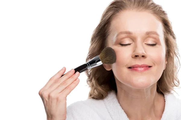 Mujer madura en albornoz usando cepillo de cosméticos mientras se aplica maquillaje aislado en blanco — Stock Photo