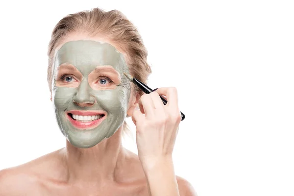 Femme mûre souriante Application masque d'argile avec pinceau cosmétique isolé sur blanc — Photo de stock