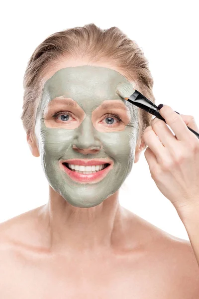 Mujer madura sonriente Aplicando máscara de arcilla con cepillo cosmético aislado en blanco - foto de stock