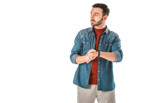 Serio uomo in denim camicie guardando altrove e toccando orologio isolato su bianco — Foto stock