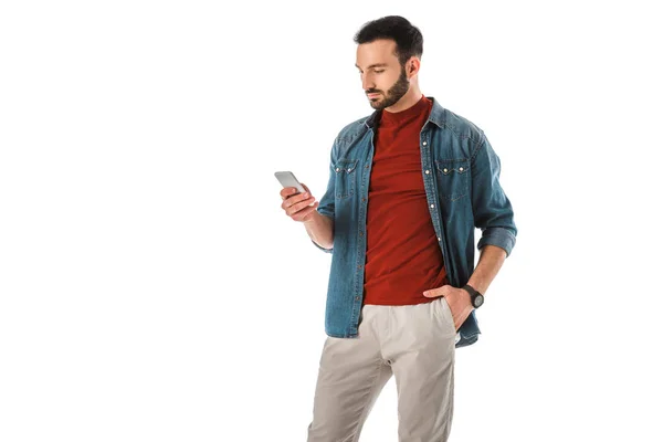 Hombre pensativo usando teléfono inteligente mientras está de pie con la mano en el bolsillo aislado en blanco - foto de stock