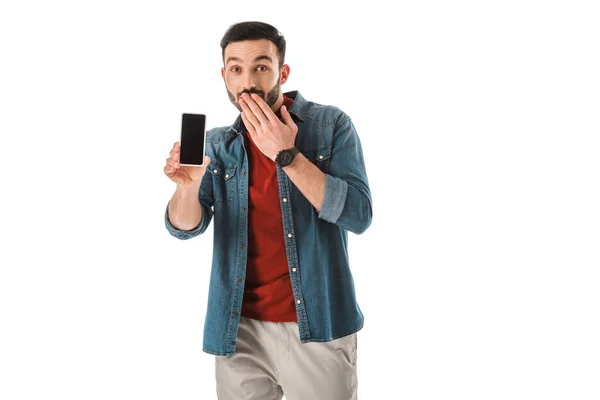 Hombre barbudo sorprendido cubriendo la boca con la mano mientras sostiene el teléfono inteligente con pantalla en blanco aislado en blanco - foto de stock