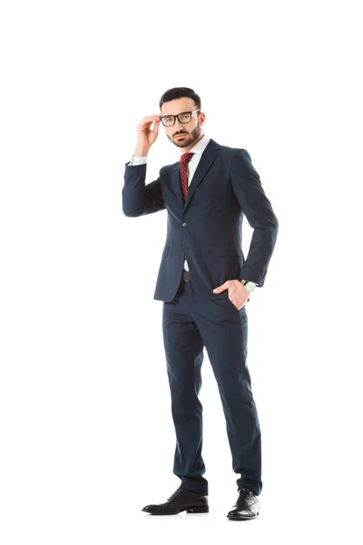 Hombre de negocios confiado de pie con la mano en el bolsillo, tocando las gafas y mirando a la cámara aislada en blanco - foto de stock