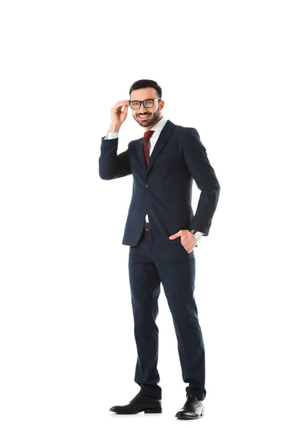 Smiling businessman standing with hand in pocket, touching glasses and looking at camera isolated on white — Stock Photo