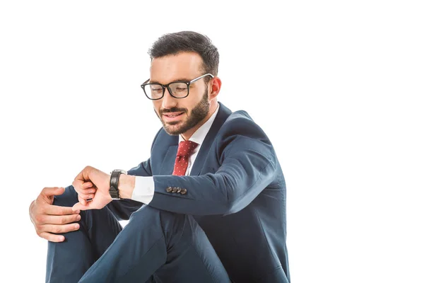 Uomo d'affari sorridente in occhiali guardando orologio mentre seduto isolato su bianco — Foto stock