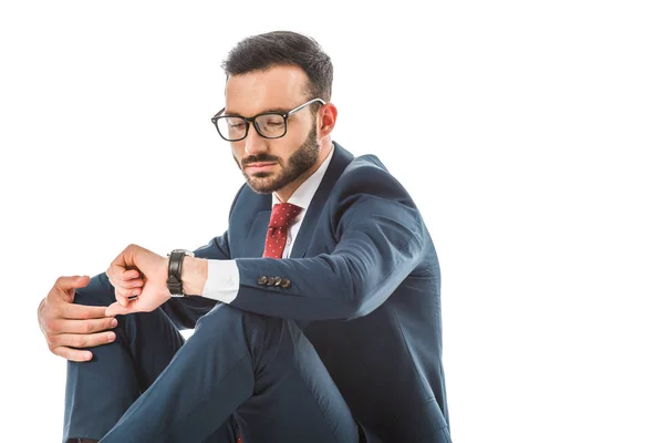 Sérieux homme d'affaires dans des lunettes regardant la montre tout en étant assis isolé sur blanc — Photo de stock