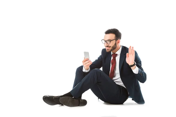 Sonriente hombre de negocios que tiene chat de vídeo en el teléfono inteligente y agitar la mano mientras está sentado en el fondo blanco - foto de stock