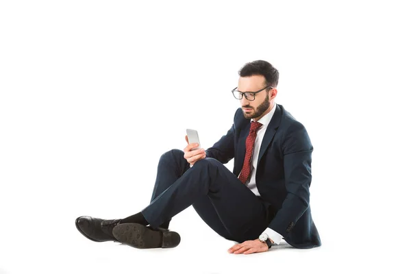 Задумчивый бизнесмен в очках, используя смартфон, сидя на белом фоне — стоковое фото