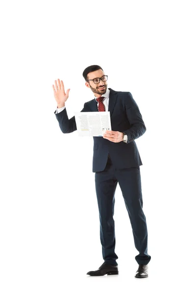 Sonriente hombre de negocios con periódico mirando hacia atrás y mostrando un gesto de saludo aislado en blanco - foto de stock
