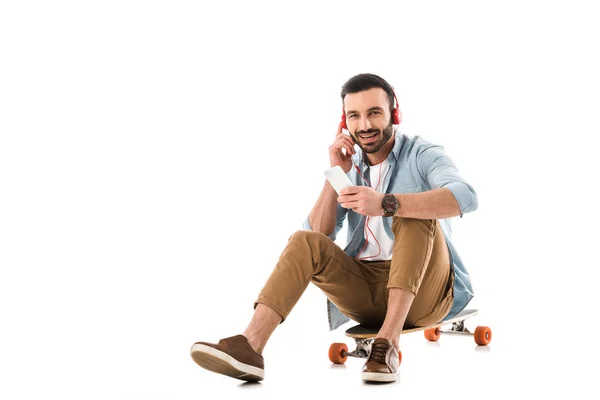 Cheerful man in headphones talking on smartphone while sitting on longboard isolated on white — Stock Photo