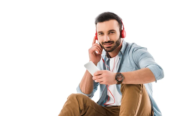 Bel homme souriant dans les écouteurs à l'aide d'un smartphone et en regardant la caméra isolée sur blanc — Photo de stock