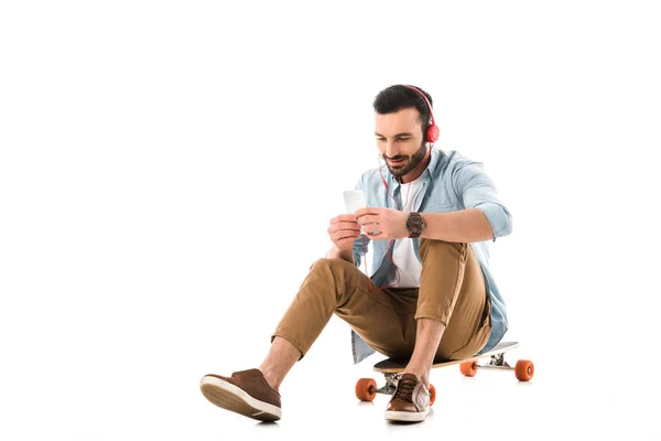 Bell'uomo sorridente che ascolta musica in cuffia mentre è seduto sul longboard e utilizza smartphone isolato su bianco — Foto stock