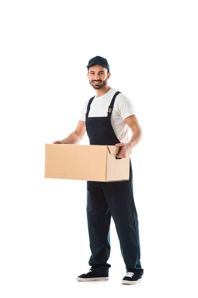 Усміхнений чоловік доставки в комбінезоні тримає картонну коробку і дивиться на камеру ізольовано на білому — стокове фото