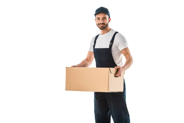 Guapo repartidor en overoles sosteniendo la caja de cartón y sonriendo a la cámara aislada en blanco - foto de stock