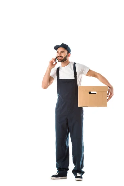 Alegre repartidor hombre en overoles sosteniendo caja de cartón y hablando en teléfono inteligente aislado en blanco - foto de stock