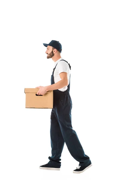 Вид збоку усміхненого чоловіка доставки в комбінезоні з картонною коробкою ізольовано на білому — стокове фото