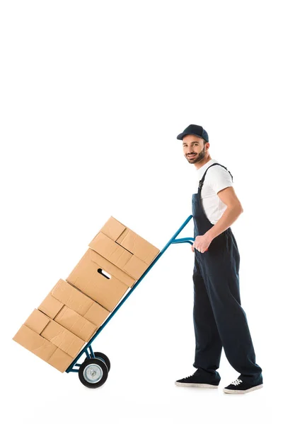 Livreur souriant transportant camion à main chargé de boîtes en carton isolé sur blanc — Photo de stock