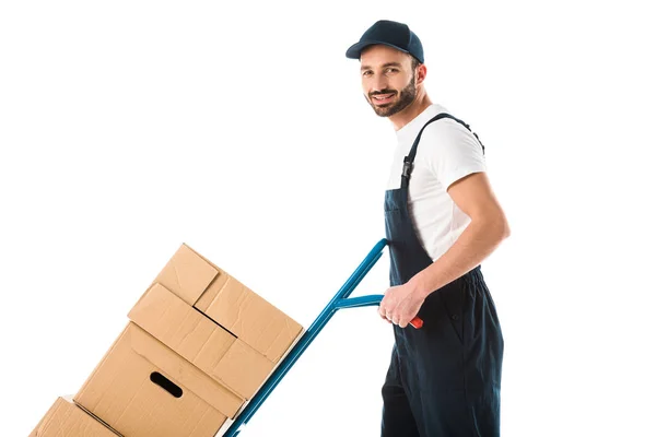 Cheerful delivery man transporting hand truck with cardboard boxes and looking at camera isolated on white — Stock Photo