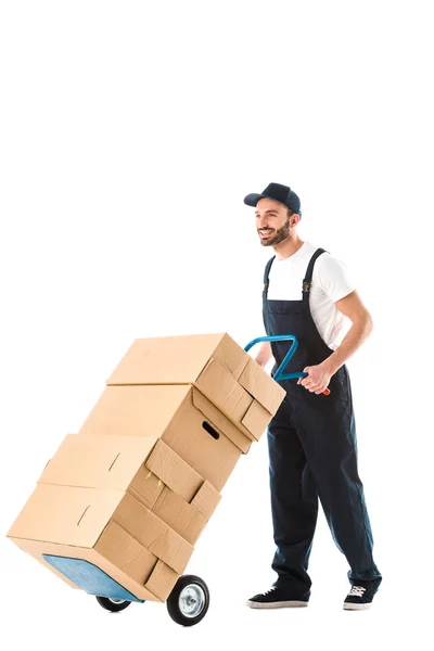 Joyeux livreur beau transportant des boîtes en carton chargé sur camion à main isolé sur blanc — Photo de stock